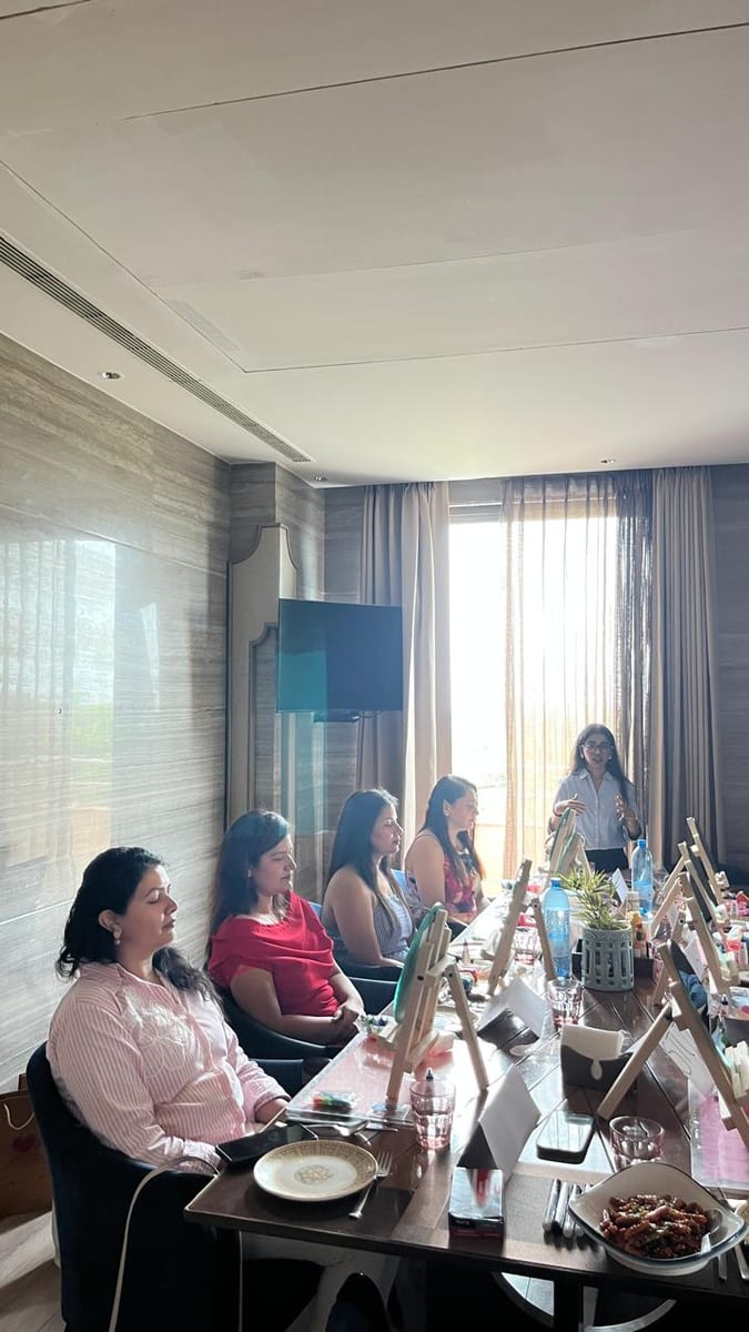 Colleagues seated around a boardroom table with mini easels and craft kits at their places, a Heart Arts India facilitator leading the session from the head of the table