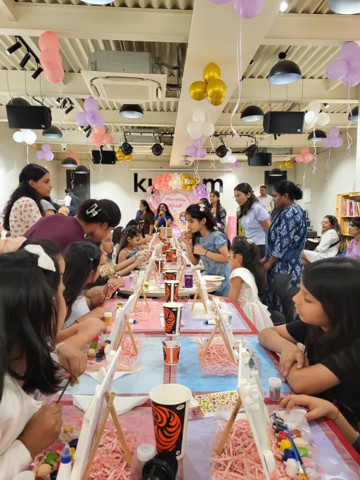 Children at a birthday celebration holding up the photo frames they decorated, balloons overhead and a Heart Arts India facilitator helping alongside them