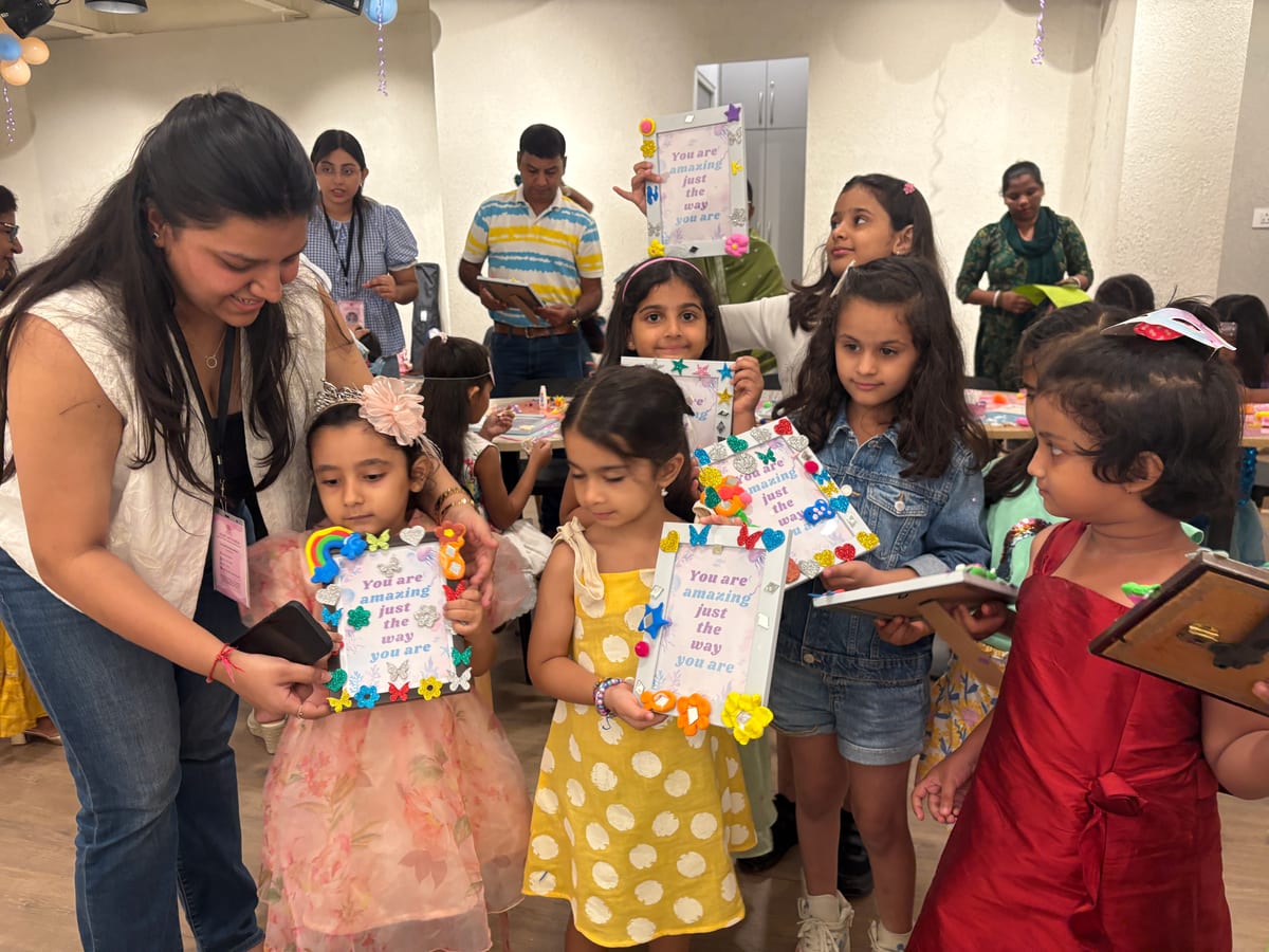 Children at a birthday celebration holding up the photo frames they decorated, balloons overhead and a Heart Arts India facilitator helping alongside them
