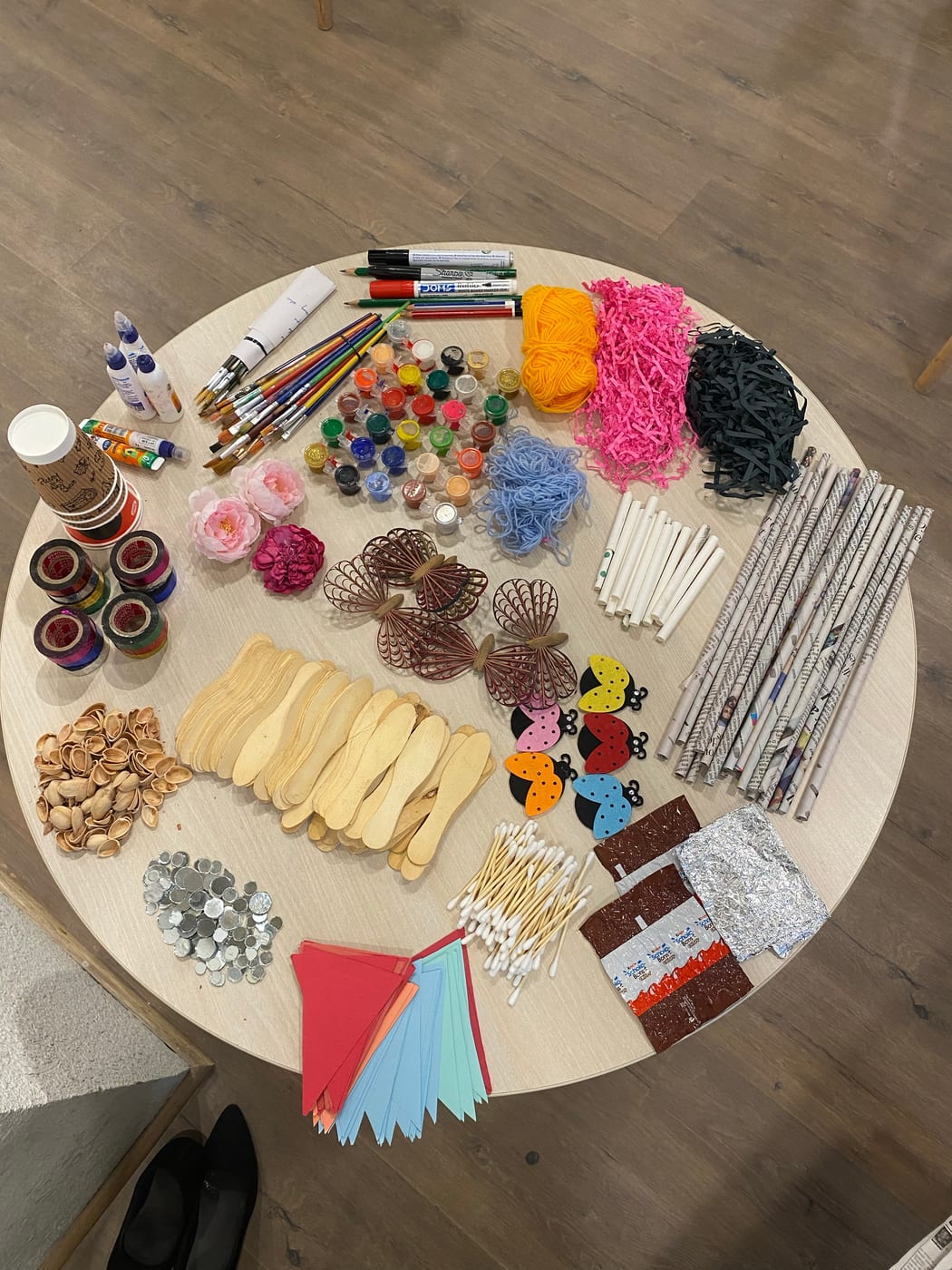 Art materials laid out on a round table before a Heart Arts India workshop — brushes, paint pots, yarn, pom-poms, beads and coloured paper.