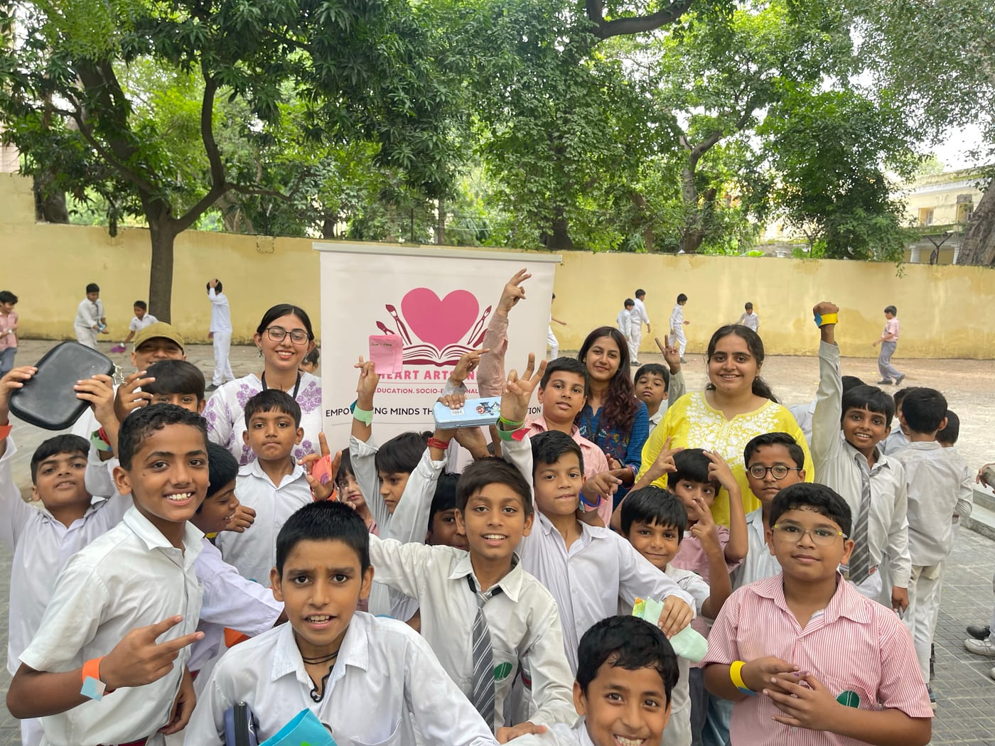 School children in uniform with their teachers on a school ground, beside the Heart Arts India banner.