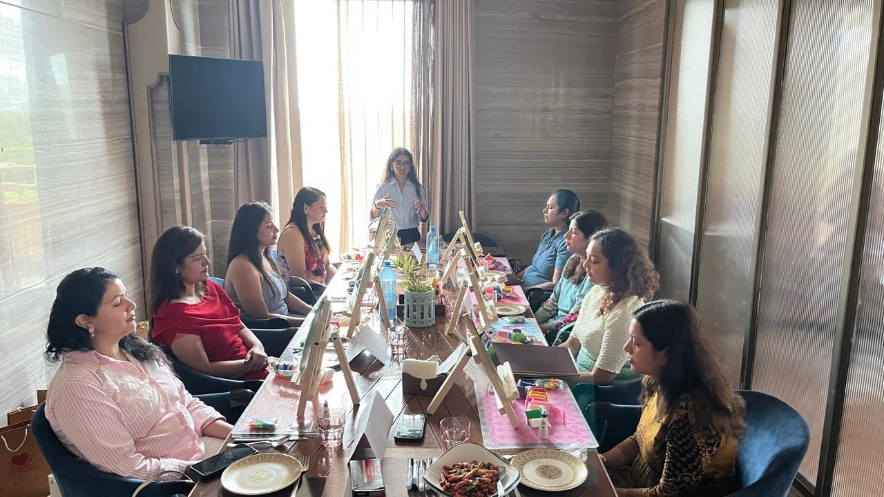 Women seated around a long table, each with a small canvas on an easel in front of them, listening to a Heart Arts India facilitator lead the session.